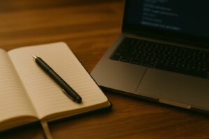 机の上にノートとペン、隣にノートPCの画面が光っている。 　アナログとデジタルが共存する絵。